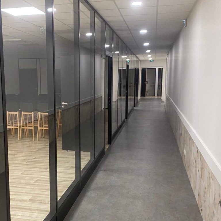 Couloir moderne avec murs en verre et chaises en bois au fond.